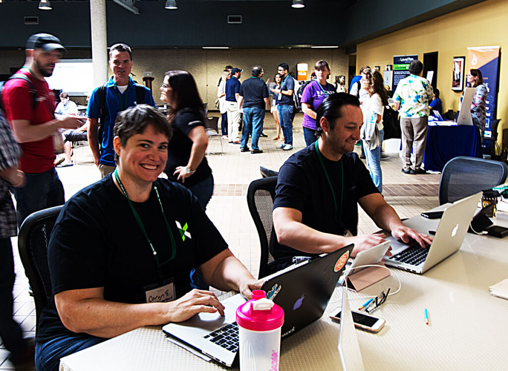 A happy woman and man using laptops at a table, surrounded by happy, chatting camp attendees.