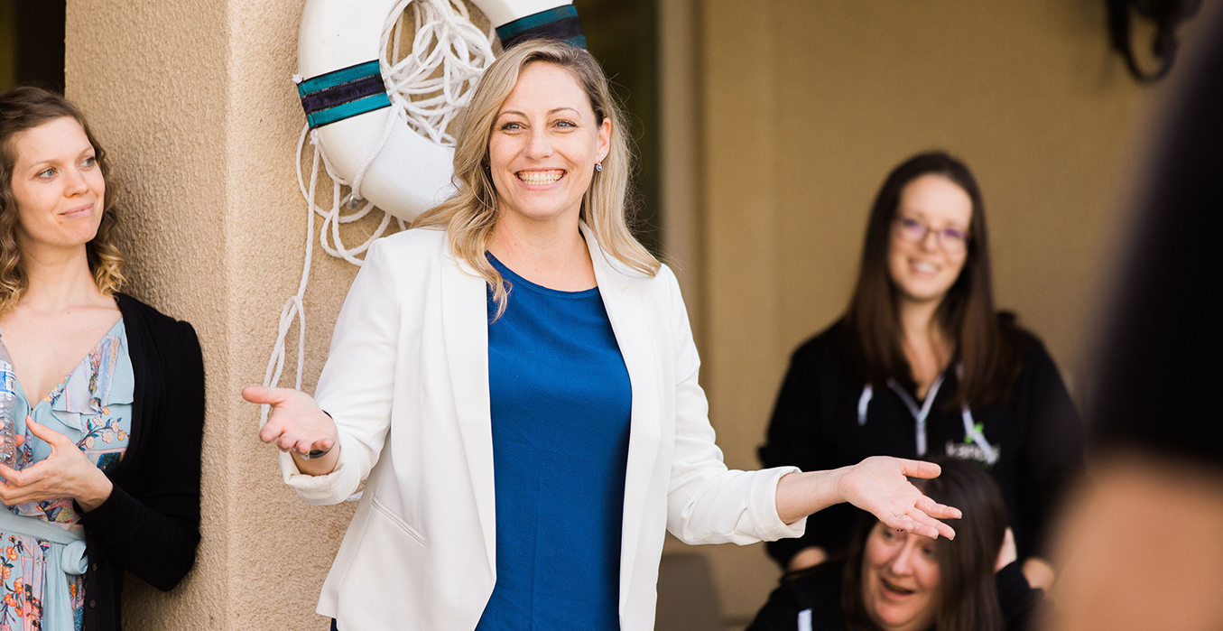 Anne Stefanyk smiling, standing in a white blazer with blue shirt, her arms outstretched.