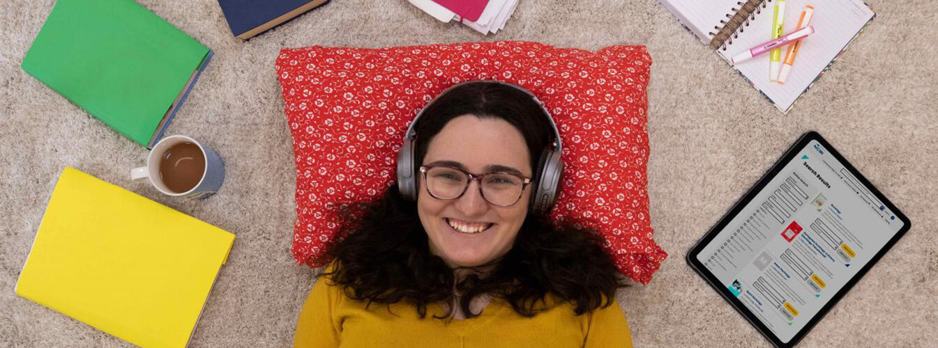 Woman shot from overhead. She's lying on her back smiling and wearing headphones, surrounded by books and a cup of coffee