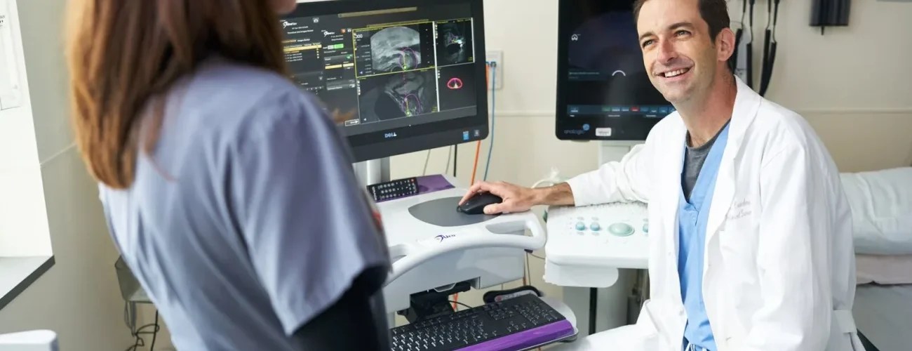 A male doctor seated at a table with a monitor showing medical images, looking up and smiling at a female doctor.