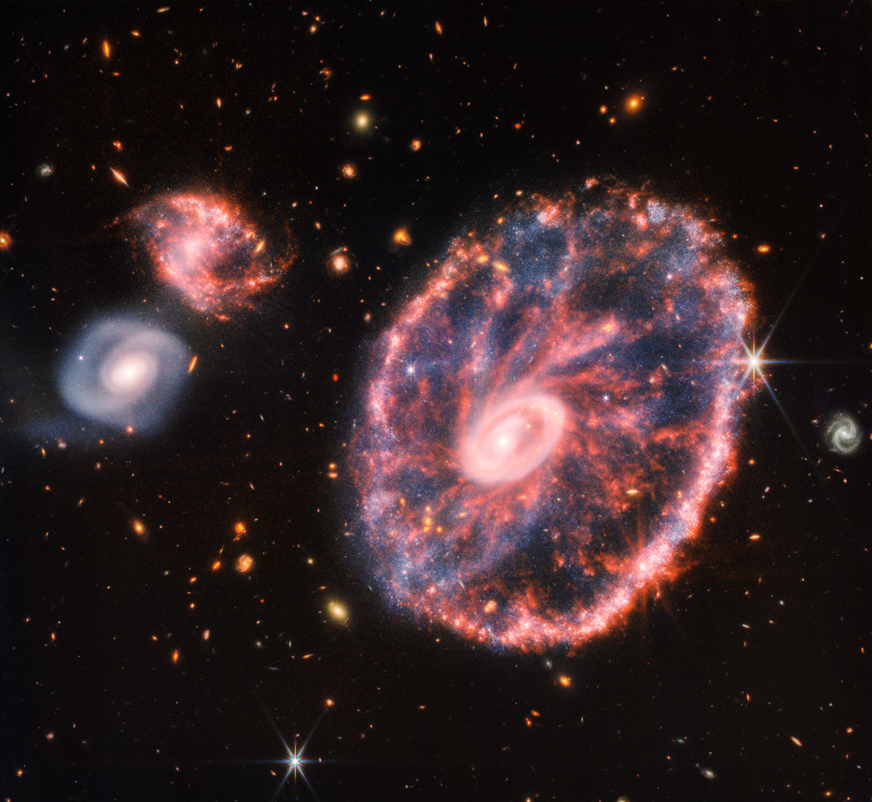 A large pink, speckled galaxy resembling a wheel with with a small, inner oval, with dusty blue in between on the right, with two smaller spiral galaxies about the same size to the left against a black background.