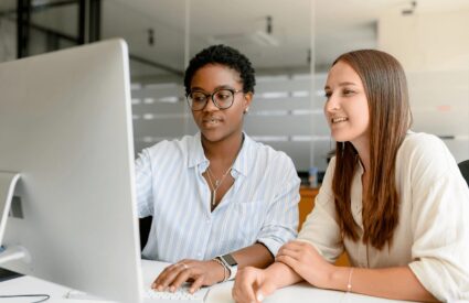 Two young women look at a desktop computer screen in an office