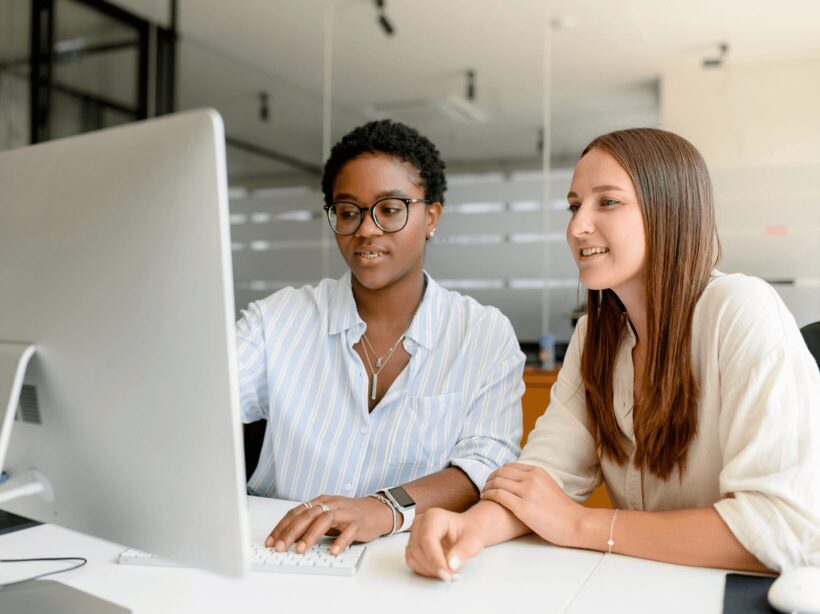 Two young women look at a desktop computer screen in an office