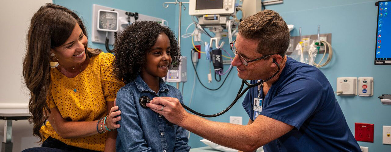 A young girl getting her heart checked by a doctor via a stethoscope as her mother looks on.