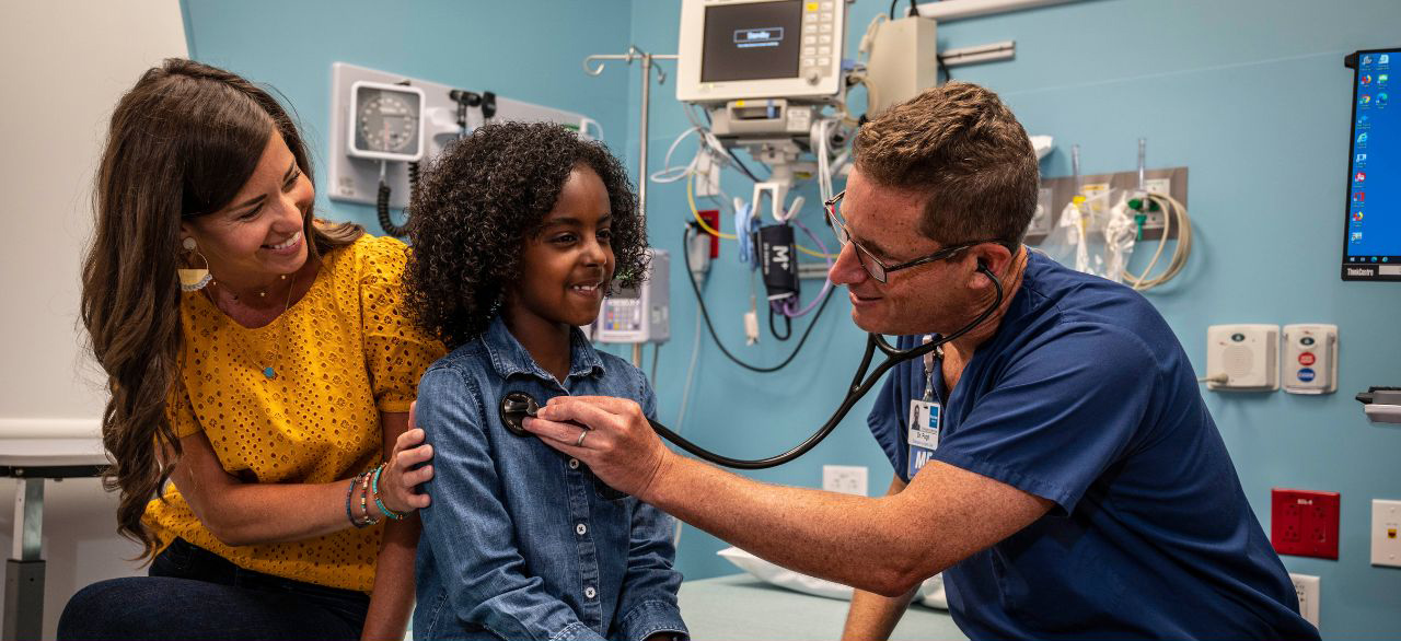 A young girl getting her heart checked by a doctor via a stethoscope as her mother looks on.