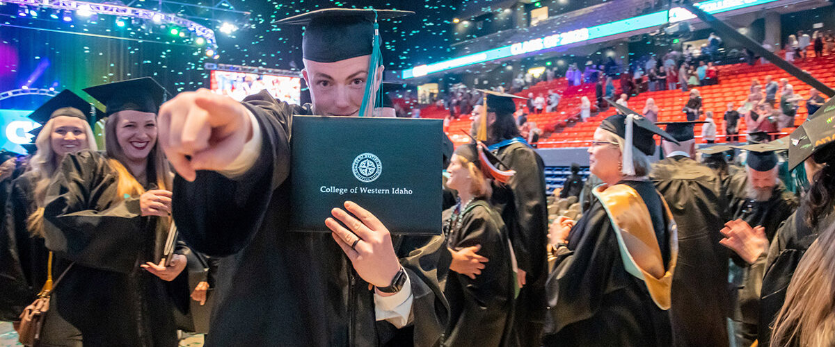 A graduate of College of Western Idaho wears his cap and gown and holds up his diploma with the school logo on it and points at the camera while confetti falls down in the stadium behind him
