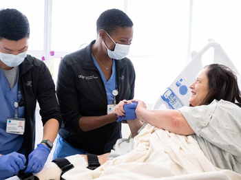 Two medical professionals in medical masks work around a female patient in a bed. The one medical professional is attaching tubes to the patient's leg, whil ethe other holds the patient's hand