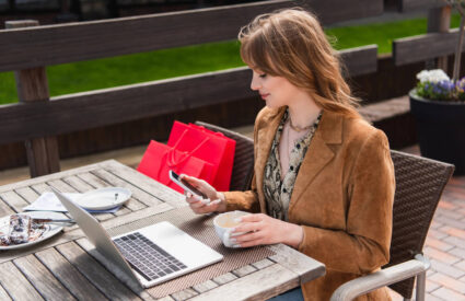 A young woman uses a laptop and mobile device while seated outside at a cafe