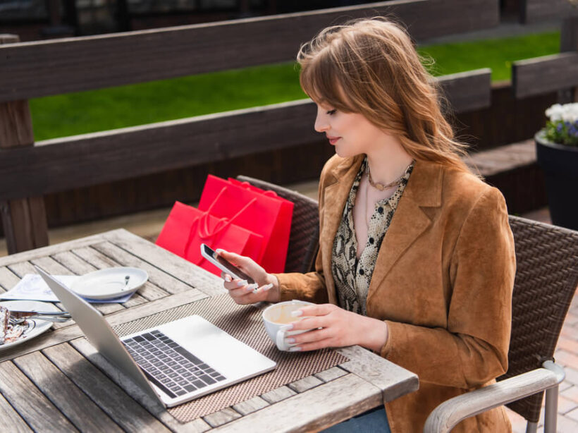 A young woman uses a laptop and mobile device while seated outside at a cafe