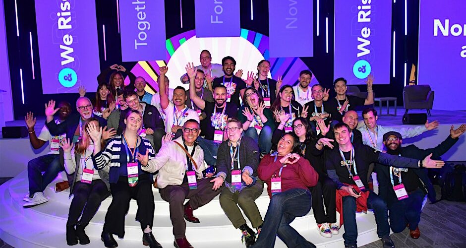 Group of about 30 people sitting on a stage smiling at an Out and Equal event, with purple banners behind them