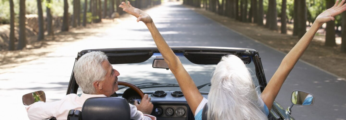 Viewed from behind, an elderly man and woman are driving away in a convertible. The woman has her arms up in the air in an expression of freedom and joy, while the man smiles at her.